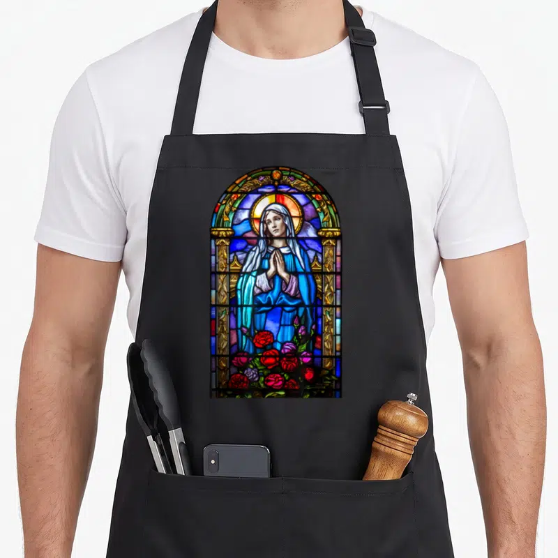 Tablier de cuisine noir avec un vitrail représentant la Sainte Vierge Marie en prière entourée de roses rouges et bleues.