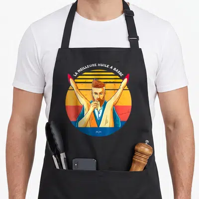Tablier de cuisine noir avec illustration d'un barbier aux jambes levées et texte La meilleure huile à barbe en arc de cercle.