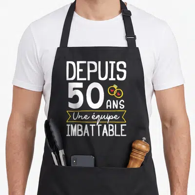 Tablier de cuisine noir avec texte blanc depuis 50 ans une équipe imbattable et deux anneaux dorés sur fond gris.