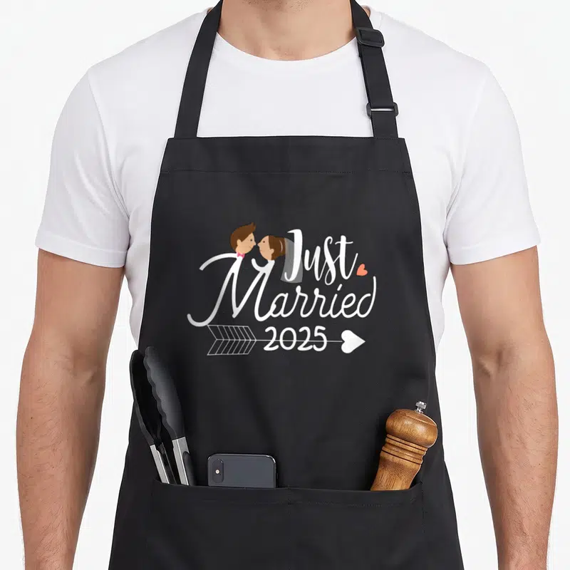 Tablier de cuisine noir avec illustration couple s'embrassant et texte Just Married 2025 traversé par une flèche.