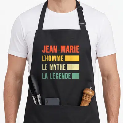 Tablier de cuisine noir avec le texte Jean-Marie l'homme le mythe la légende en lettres orange jaune et vertes.