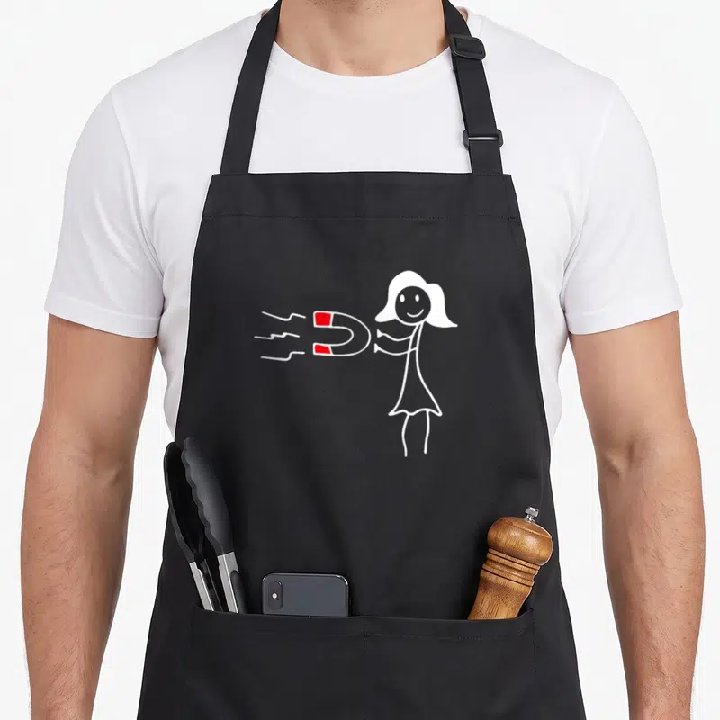 Tablier de cuisine noir avec une silhouette blanche souriante tenant un aimant rouge et blanc, sur fond gris uni.