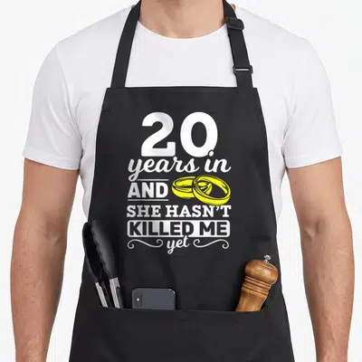 Tablier de cuisine noir avec texte blanc 20 years in and she hasn't killed me yet et deux anneaux jaunes entrelacés.