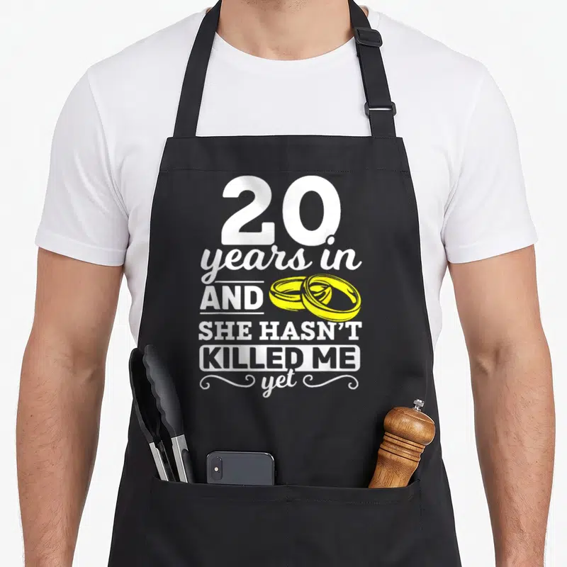 Tablier de cuisine noir avec texte blanc 20 years in and she hasn't killed me yet et deux anneaux jaunes entrelacés.