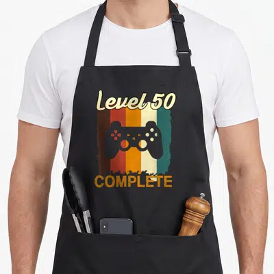 Tablier de cuisine noir avec manette de jeu vidéo et texte Level 50 Complete sur fond rayé vintage.