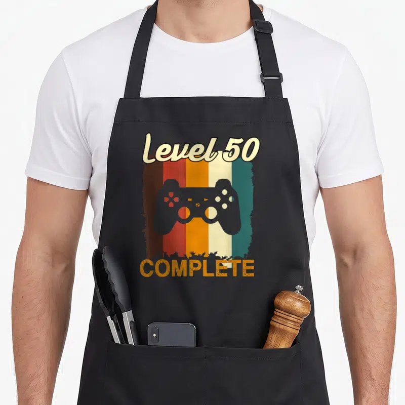 Tablier de cuisine noir avec manette de jeu vidéo et texte Level 50 Complete sur fond rayé vintage.