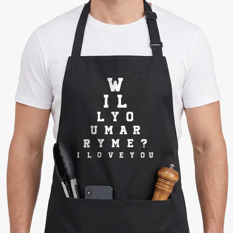 Tablier de cuisine noir avec un test oculaire blanc formant la phrase Veux-tu m'épouser et I love you en bas.