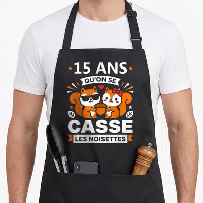 Tablier de cuisine noir avec deux écureuils orange tenant une noix et le texte 15 ans qu'on se casse les noisettes.