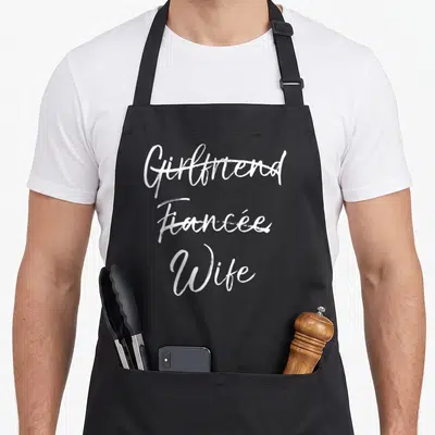 Tablier de cuisine noir avec les mots Girlfriend et Fiancée barrés au-dessus du mot Wife en calligraphie blanche.