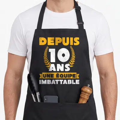 Tablier de cuisine noir avec inscription blanche et orange depuis 10 ans une équipe imbattable encadré de couronnes de laurier dorées.