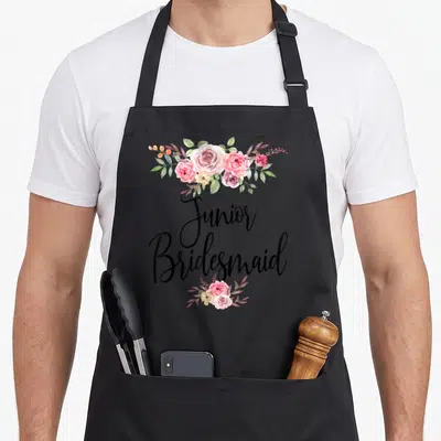 Tablier de cuisine noir avec inscription Junior Bridesmaid encadrée de roses roses et de feuillage vert.