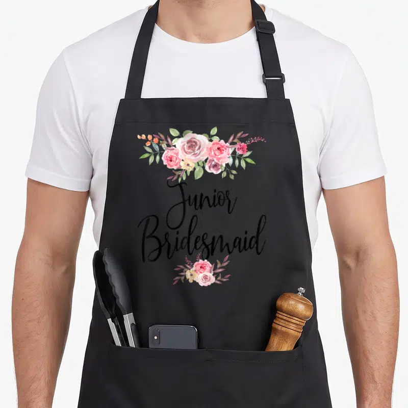Tablier de cuisine noir avec inscription Junior Bridesmaid encadrée de roses roses et de feuillage vert.