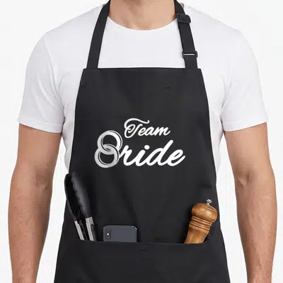 Tablier de cuisine noir avec l'inscription Team Bride et une alliance blanche stylisée en forme de S.