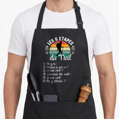 Tablier de cuisine noir avec silhouette de trailleur et texte humoristique sur les étapes de l'épreuve.