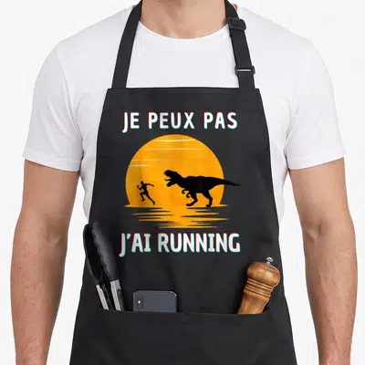 Tablier de cuisine noir avec une illustration d'un homme fuyant un dinosaure sous un soleil orange et le texte Je peux pas j'ai running.