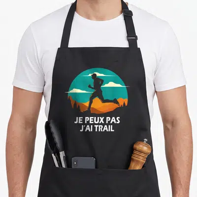 Tablier de cuisine noir avec silhouette de coureur de trail et texte Je peux pas j'ai trail en blanc sur fond de paysage stylisé.