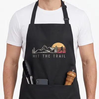 Tablier de cuisine noir avec illustration d'untrailleur, montagnes et texte Hit The Trail en style rétro.