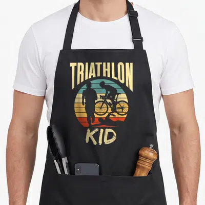 Tablier de cuisine noir avec illustration vintage Triathlon Kid montrant un coureur, un cycliste et un nageur devant un soleil multicolore.