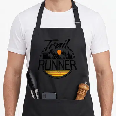 Tablier de cuisine noir avec logo Trail Runner représentant des montagnes et un soleil orange dans un style vintage circulaire.