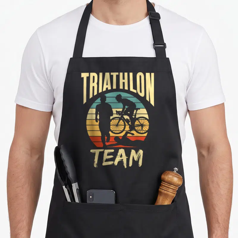 Tablier de cuisine noir avec illustration rétro Triathlon Team montrant un nageur, un cycliste et un coureur de foot sur fond de soleil couchant.