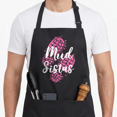 Tablier de cuisine noir avec deux grandes empreintes de bottes roses et le texte Mud Sistas en lettres blanches cursives au centre.