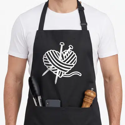 Tablier de cuisine noir avec un motif blanc représentant un coeur formé par une pelote de laine traversée par deux aiguilles.