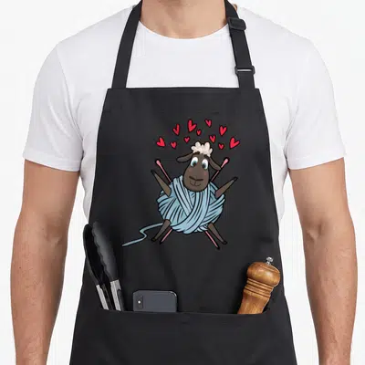 Tablier de cuisine noir avec un mouton enveloppé dans un fil bleu et des aiguilles à tricoter roses.