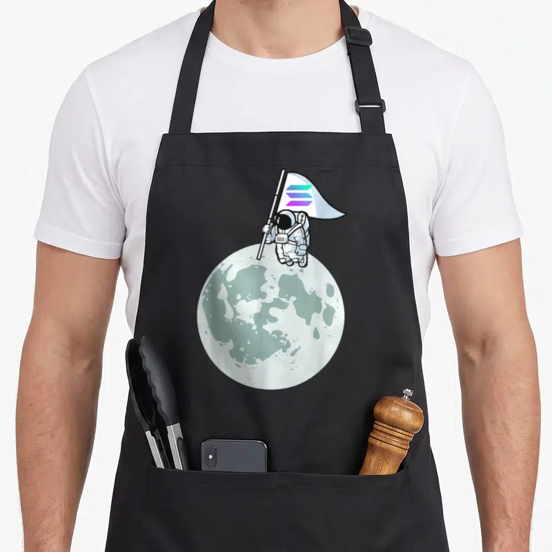 Tablier de cuisine noir avec illustration d'un astronaute plantant un drapeau aux trois bandes colorées sur une surface lunaire.