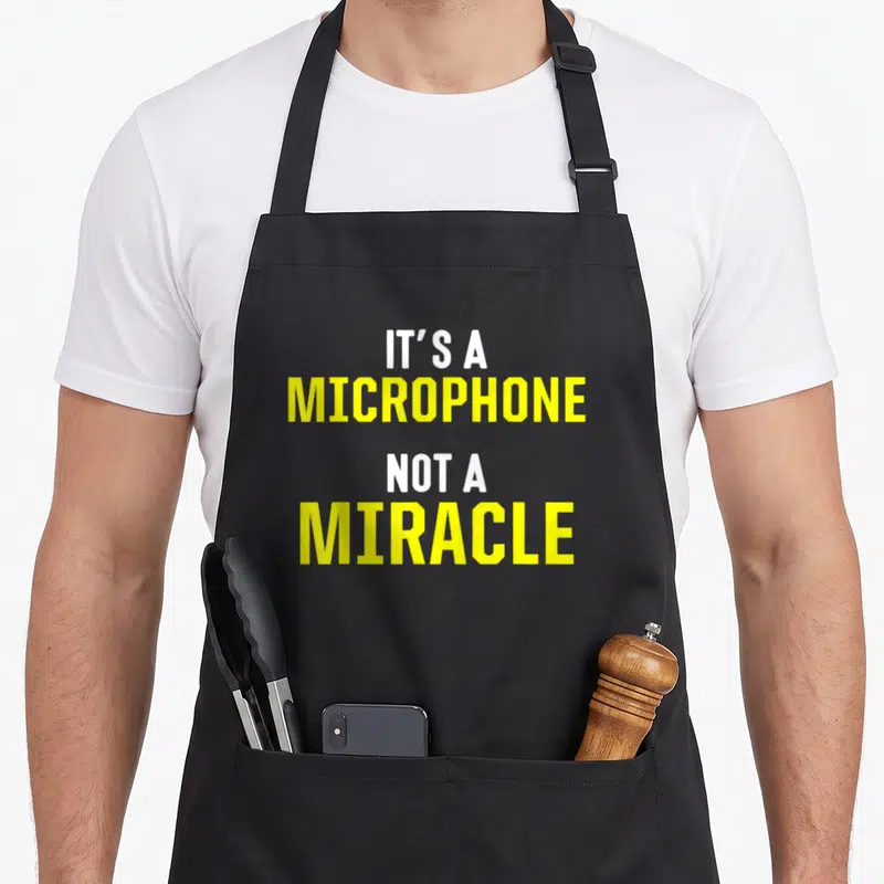 Tablier de cuisine noir avec le texte It's a microphone not a miracle en jaune et blanc.