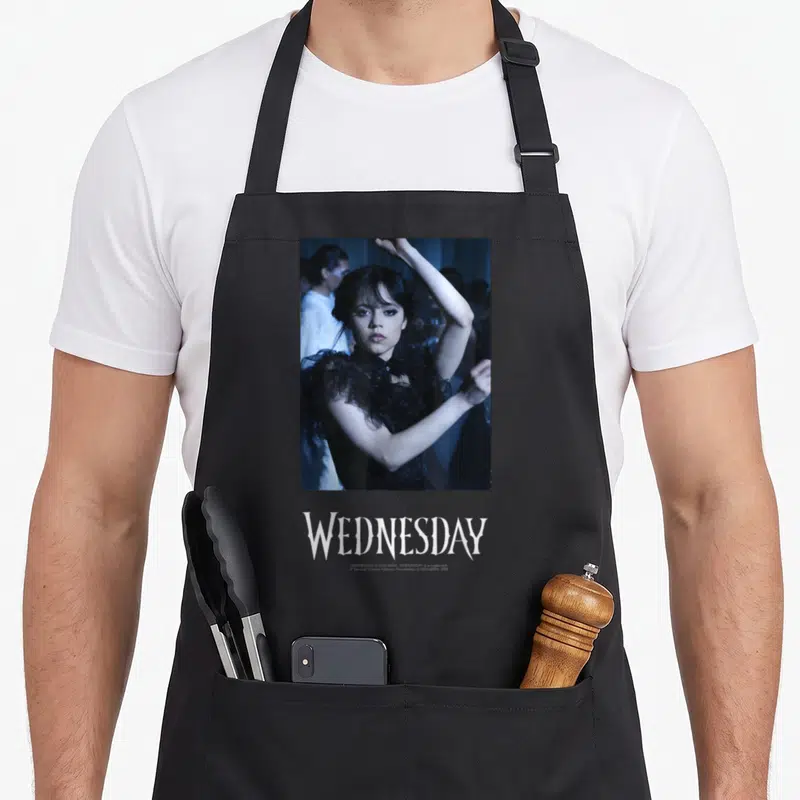 Tablier de cuisine noir avec une scène de danse et le nom WEDNESDAY imprimé en bas.