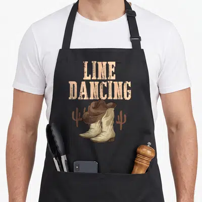 Tablier de cuisine noir avec illustration de bottes de cowboy, chapeau et texte Line Dancing en style western.