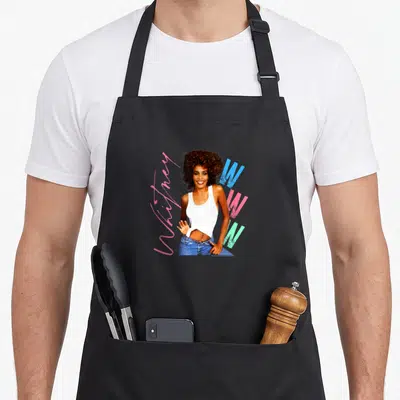 Tablier de cuisine noir avec une illustration de Whitney Houston souriante et des lettres colorées.