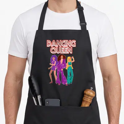 Tablier de cuisine noir avec trois femmes stylisées en tenues disco et l'inscription Dancing Queen en lettres rétro.