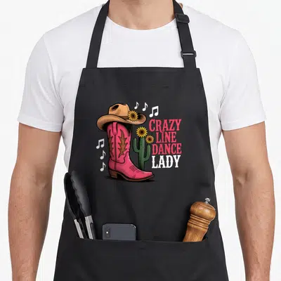 Tablier de cuisine noir avec une botte rose, un chapeau cowboy, des cactus et le texte CRAZY LINE DANCE LADY imprimé.