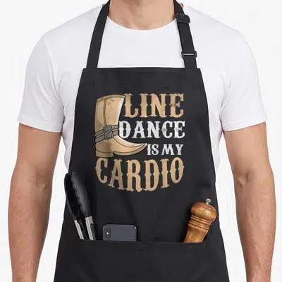 Tablier de cuisine noir avec une bottine western et le texte Line Dance is My Cardio imprimé en beige et blanc.