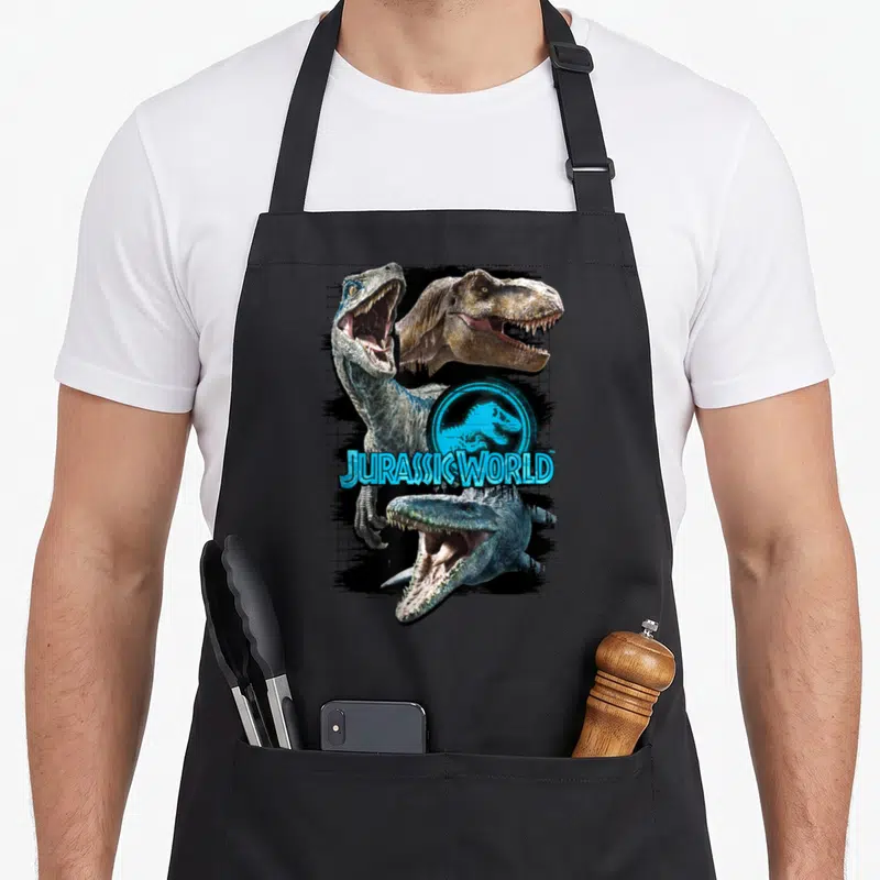 Tablier de cuisine noir avec illustration de dinosaures et logo Jurassic World bleu cyan sur fond quadrillé.