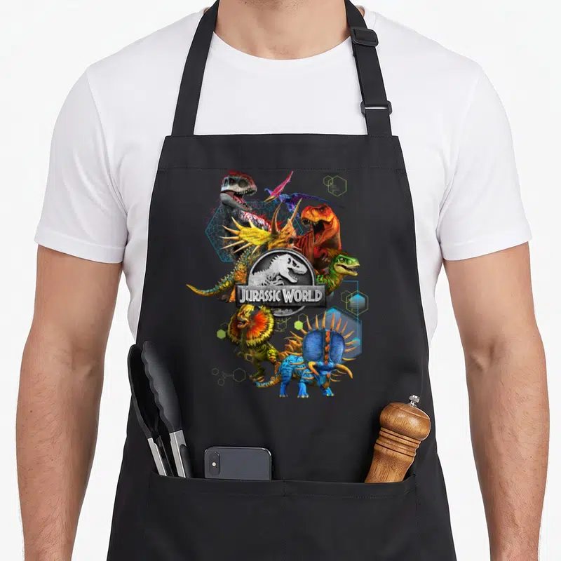 Tablier de cuisine noir avec un collage de dinosaures colorés et le logo Jurassic World au centre.