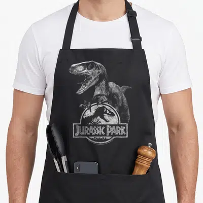 Tablier de cuisine noir avec illustration d'un Velociraptor tenant le logo Jurassic Park en blanc usé.