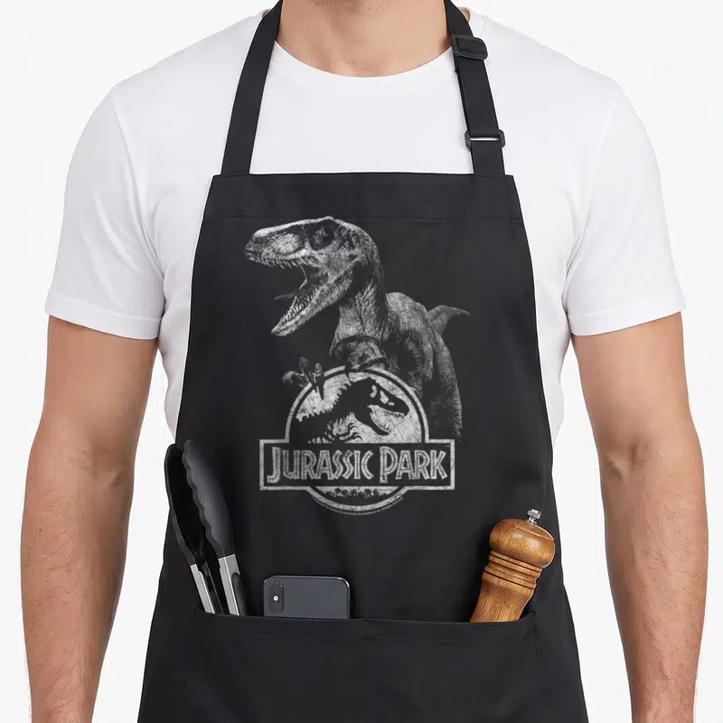Tablier de cuisine noir avec illustration d'un Velociraptor tenant le logo Jurassic Park en blanc usé.