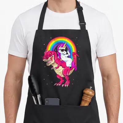 Tablier de cuisine noir avec une licorne blanche chevauchant un t rex rose sous un arc en ciel coloré.