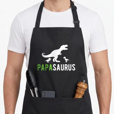 Tablier de cuisine noir avec silhouette T-Rex blanc, deux petits dinosaures et le mot Papasaurus en vert et blanc.
