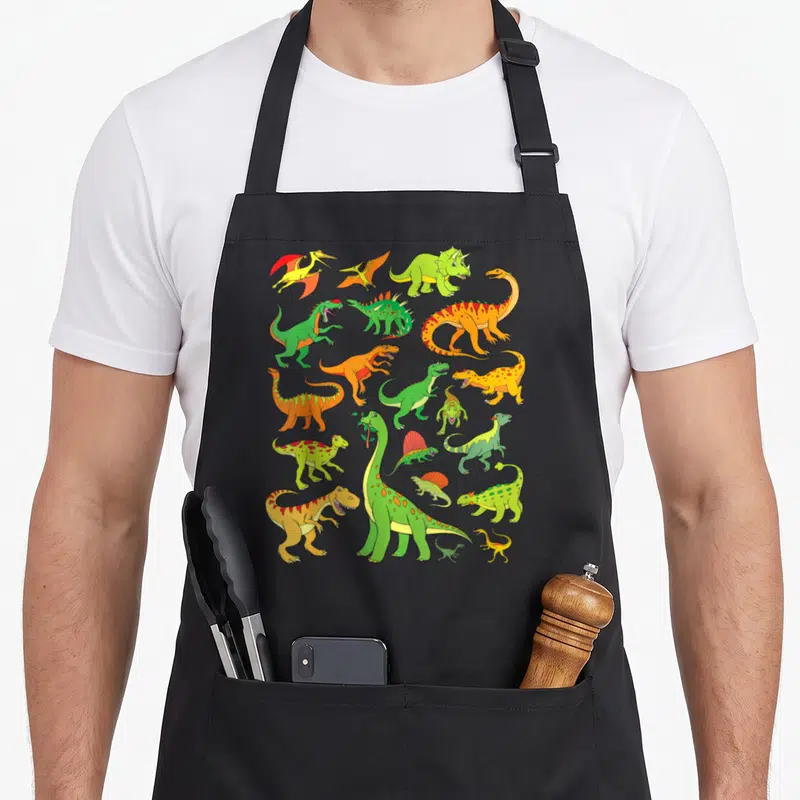 Tablier de cuisine noir avec une illustration regroupant divers dinosaures verts, oranges et jaunes sur fond uni.