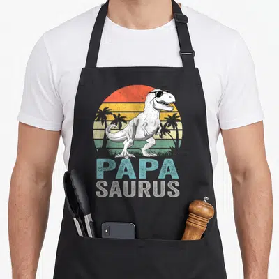 Tablier de cuisine noir avec un T Rex en lunettes de soleil et le texte Papa Saurus sur fond de soleil rétro.