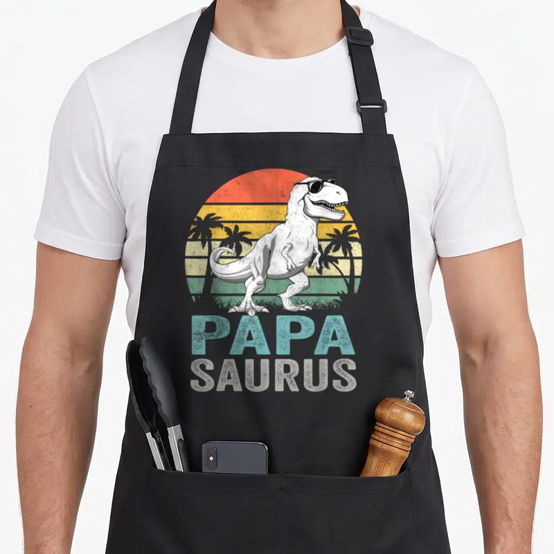 Tablier de cuisine noir avec un T Rex en lunettes de soleil et le texte Papa Saurus sur fond de soleil rétro.