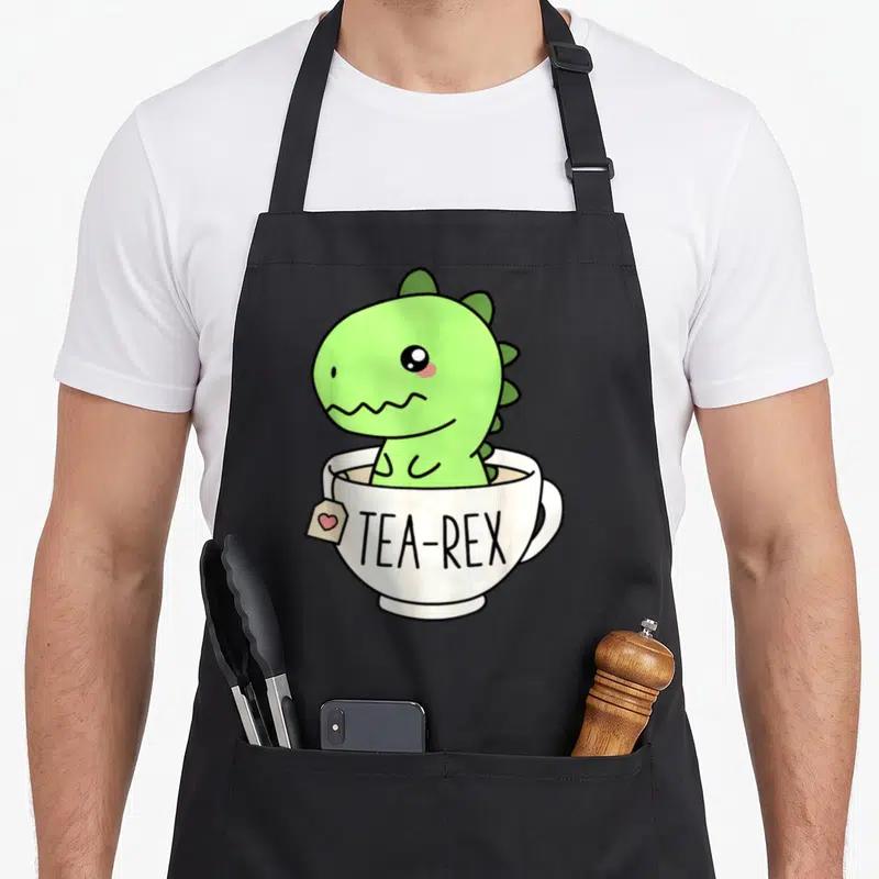 Tablier de cuisine noir avec un dinosaure vert kawaii assis dans une tasse blanche portant l'inscription Tea-Rex.