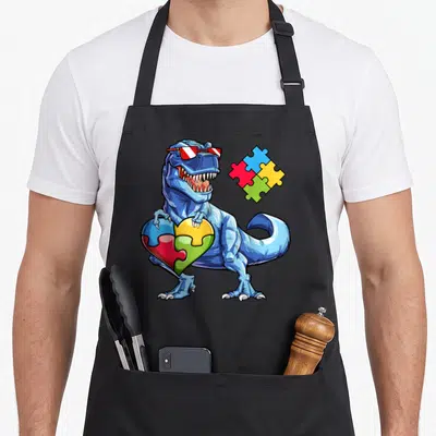 Tablier de cuisine noir avec dinosaure bleu tenant un cœur en puzzle multicolore et portant des lunettes de soleil rouges.