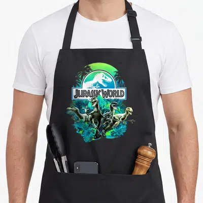 Tablier de cuisine noir avec illustration de trois raptors dans une jungle verte et bleue sous un logo blanc.