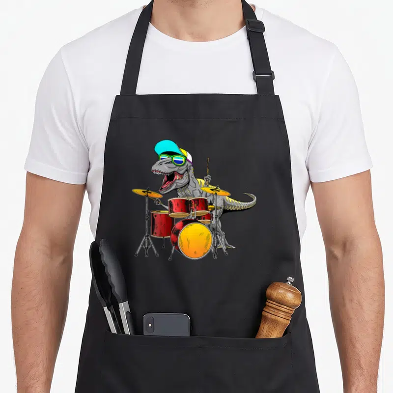 Tablier de cuisine noir avec illustration d'un T rex batteur portant une casquette et des lunettes de soleil devant une batterie rouge.