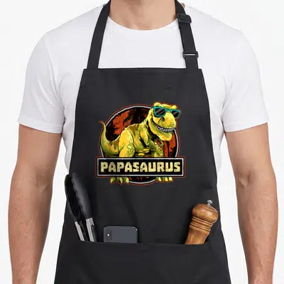 Tablier de cuisine noir avec un Tyrannosaure aux lunettes de soleil et l'inscription PAPASaurus en lettres jaunes.