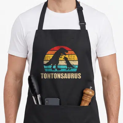 Tablier de cuisine noir avec silhouette de deux dinosaures et le mot Tontonsaurus en lettres capitales beiges sous un soleil rayé.